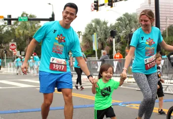 Outdoor running race with a family of three smiling at the finish line. The tall dad wears all blue, holding hands with his toddle and mom. Mom is wearing a matching Turkey Gobble and pushing the empty stroller.
