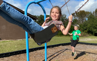 two kids on outdoor swings. girl wearing jeans in the center image, boy wearing green shirt swings in background.