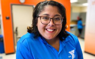 Headshot of female staff smiling at the camera in the YMCA Welcome Center. Staff is wearing a blue polo shirt with a Tampa Y logo, orange wall in background.